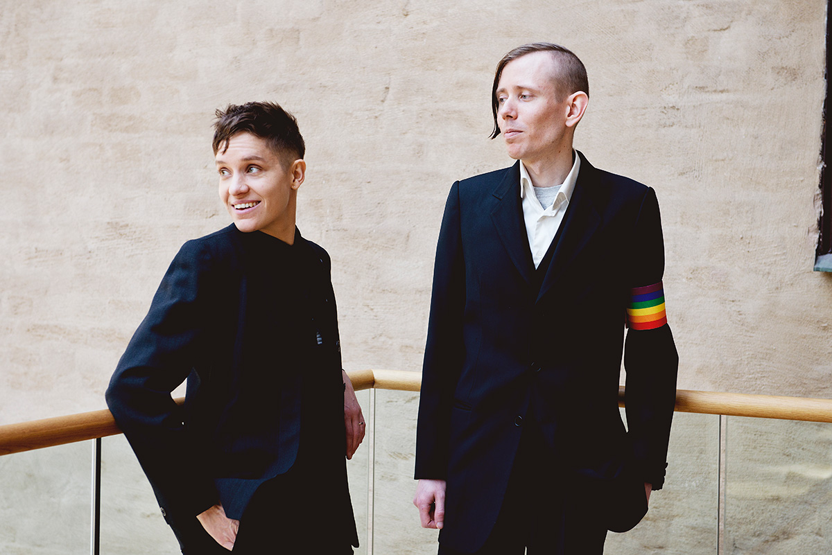 Two people dressed i black suits with a rainbow pride band around wrapped their arm